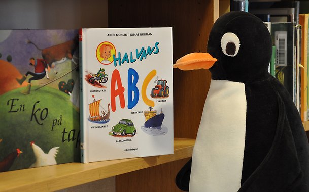Foto av gosedjur (pingvin) som sitter och tittar in i en bokhylla där det står två stycken bilderböcker.