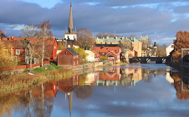 Höstvy över Arbogaån. Gråblått spegelblankt vatten med äldre faluröda byggnader på vardera sidan om vattnet.
